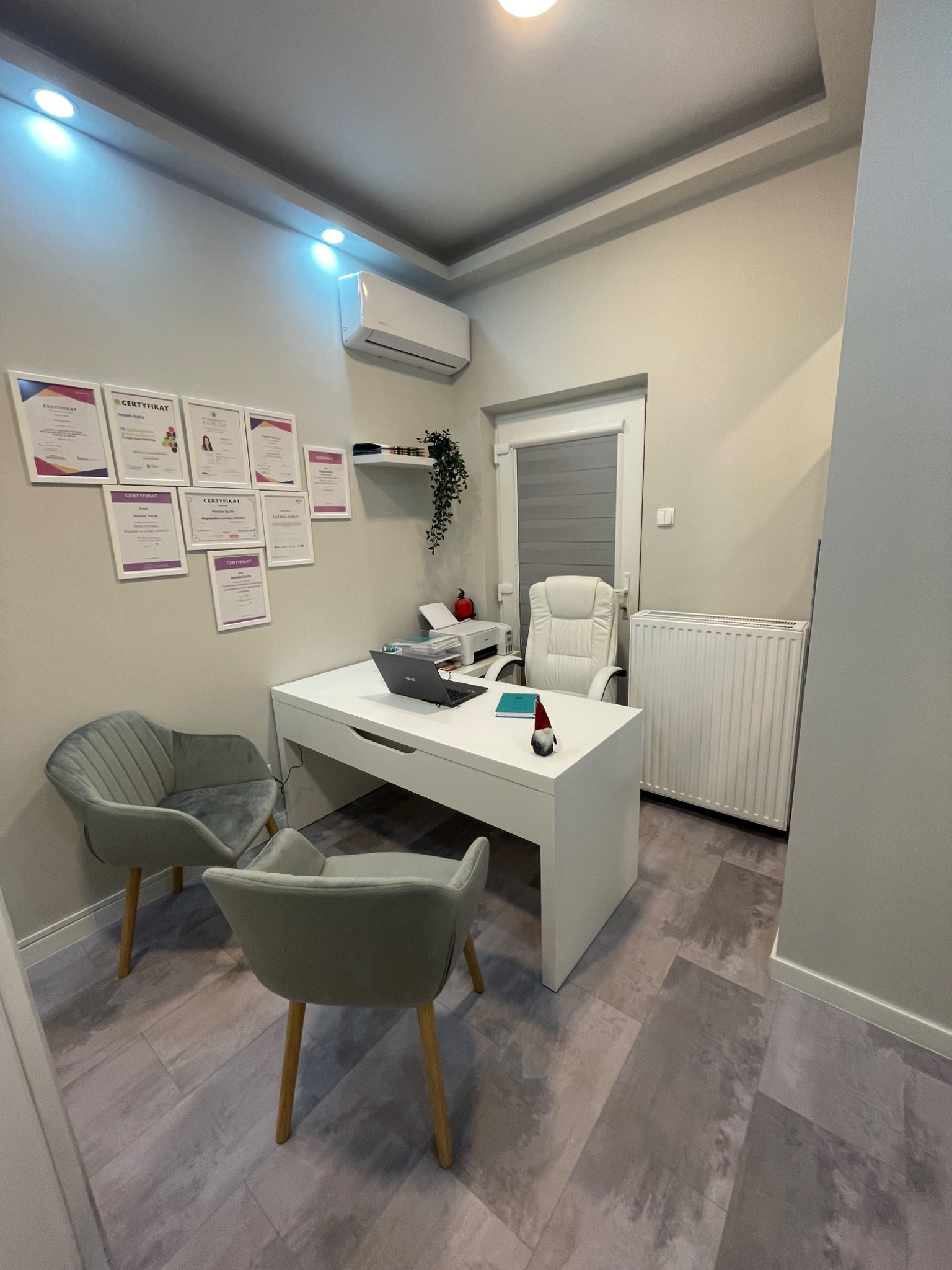 Jasne wnętrze gabinetu z biurkiem, krzesłami dla pacjentów i ścianą z certyfikatami.