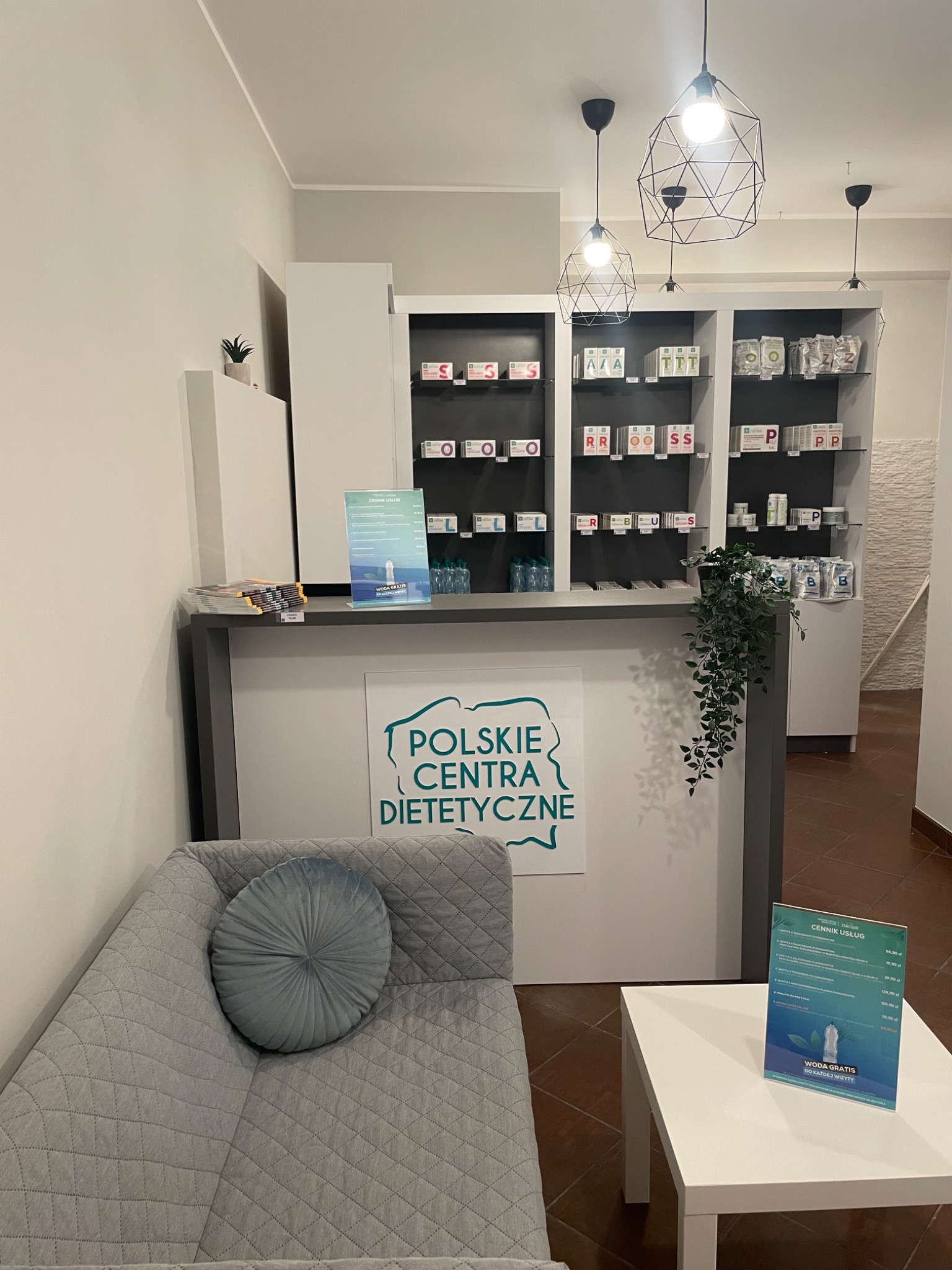 Wnętrze gabinetu z ladą recepcyjną, sofą z okrągłą poduszką, stolikiem z cennikiem i regałem z suplementami diety w tle, oświetlone wiszącymi lampami o geometrycznych kloszach.