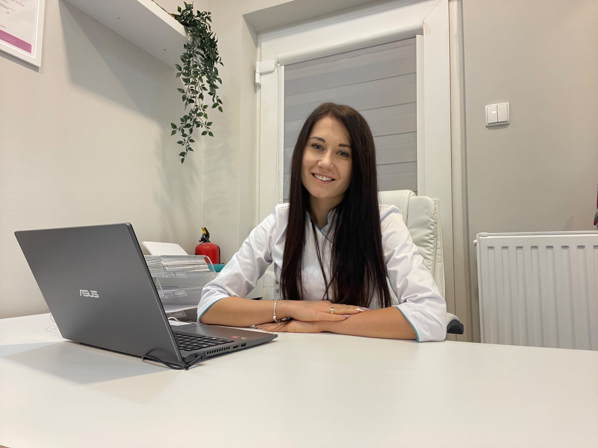Uśmiechnięta kobieta w białym uniformie siedzi za biurkiem z laptopem Asus w jasnym gabinecie z roletami i rośliną zwisającą z półki.