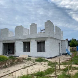 Wznoszenie ścian parterowego domu z białych bloczków, widoczna konstrukcja zbrojeniowa na gruncie i przenośna toaleta w tle.