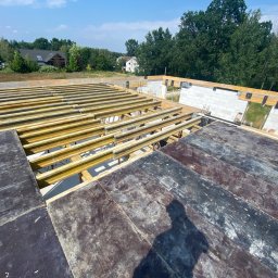 Bohdan Kostiuchyk Dream House - Wysoki kąt widzenia na fundamenty domu w trakcie budowy, widoczne szalunki i zbrojenie, tło z zielenią i domami.