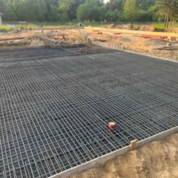 Siatka zbrojeniowa ułożona w szalunku fundamentowym, widoczne wystające rury kanalizacyjne, w tle dom w budowie i teren budowy z wykopami.