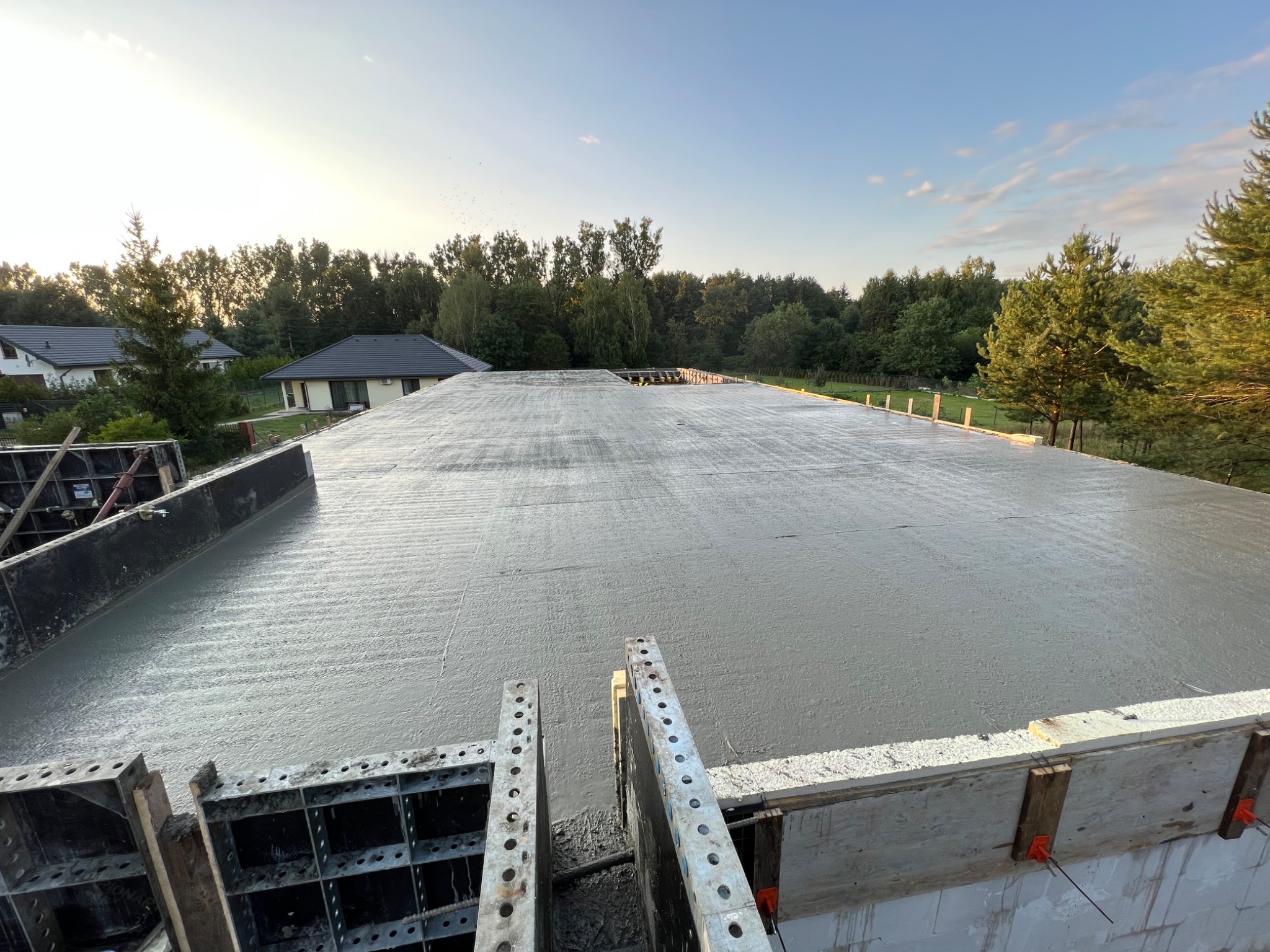 Wylany, świeży beton na stropie budynku w trakcie budowy, widoczne szalunki i zbrojenia, w tle zabudowa jednorodzinna i drzewa.