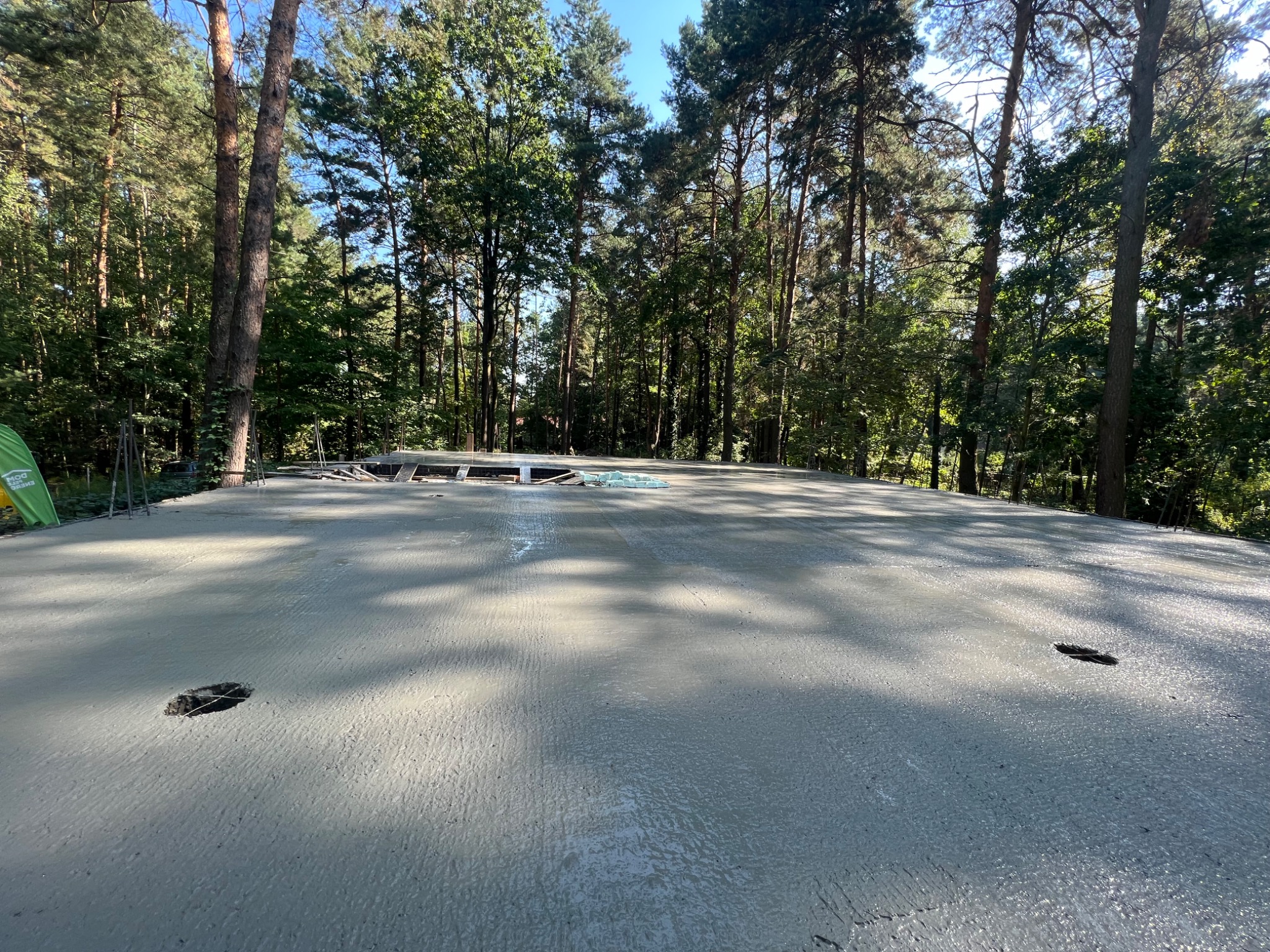 Świeżo wylana, gładka płyta fundamentowa z widocznymi otworami na instalacje, otoczona lasem iglastym w słoneczny dzień.