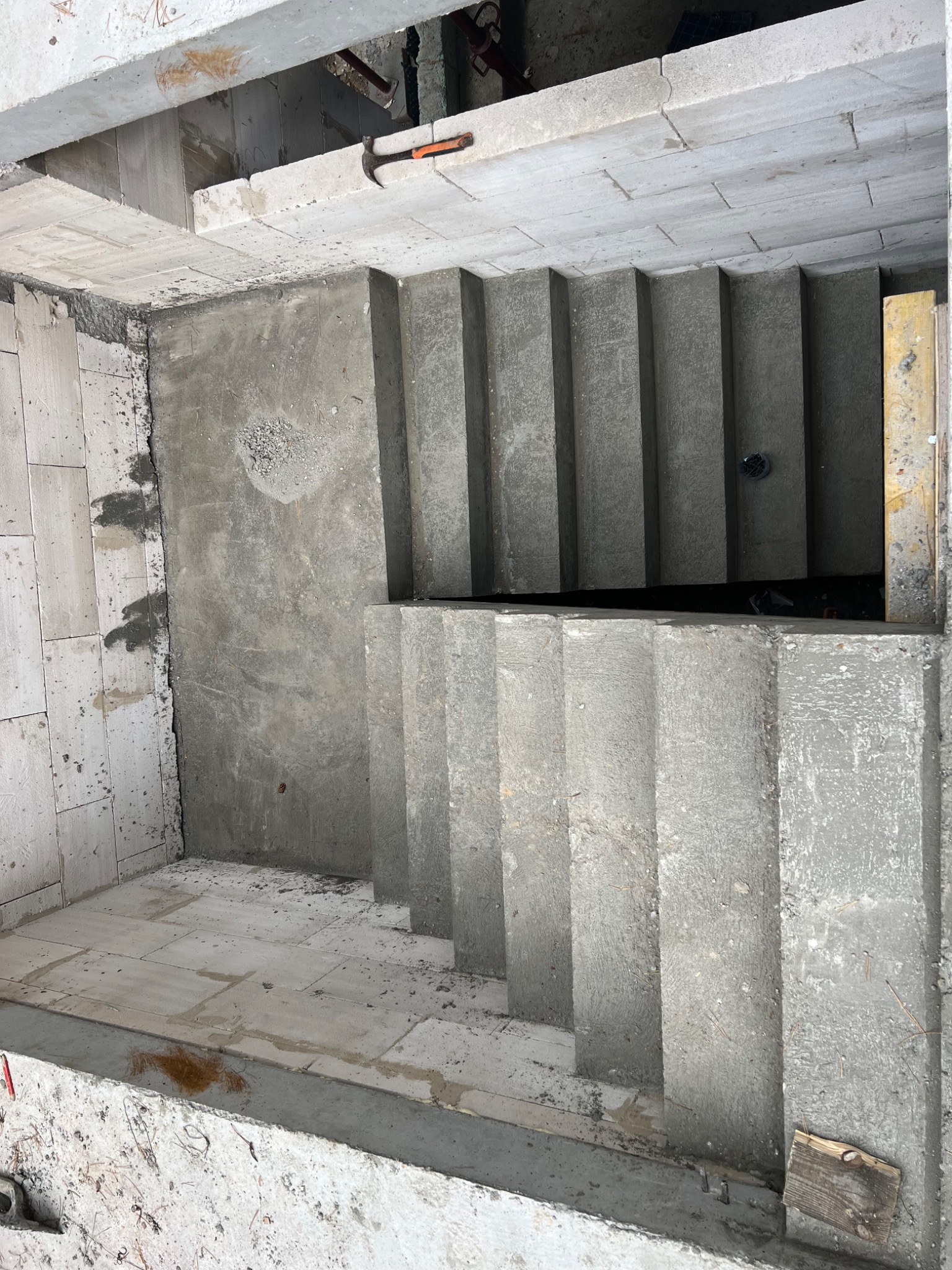 Wykop pod schody w surowym stanie z widocznym betonem, cegłami i narzędziem (młotek) na górnej krawędzi.
