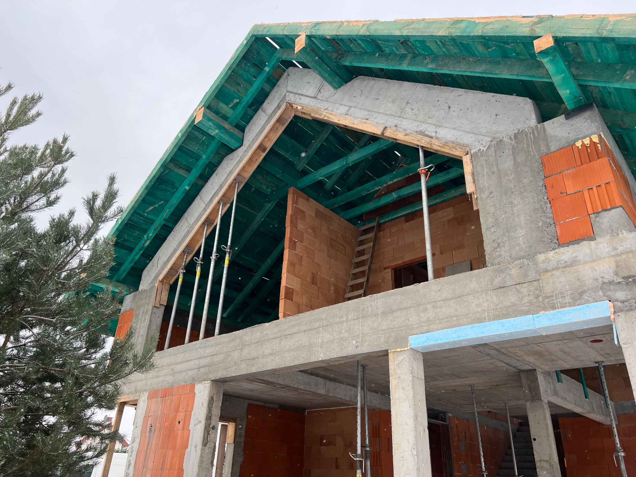 Wznoszenie dwukondygnacyjnego domu z betonu i cegły, widoczne szalunki, podpory, drewniana drabina i zielona więźba dachowa w trakcie budowy.