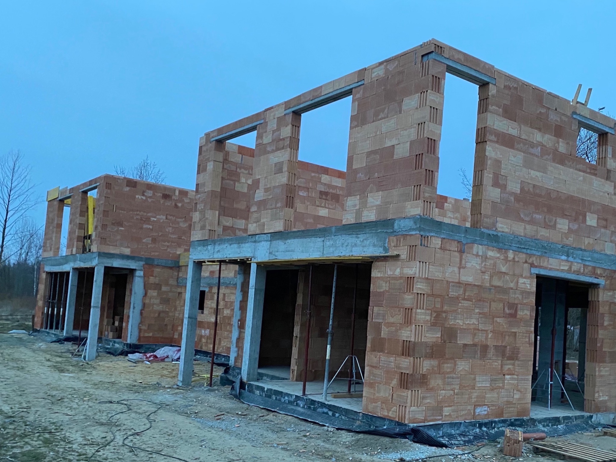 Dwa budynki w stanie surowym otwartym, wzniesione z czerwonej cegły, widoczne stalowe nadproża okienne, szalunki na stropie, teren budowy z piaskiem i folią.