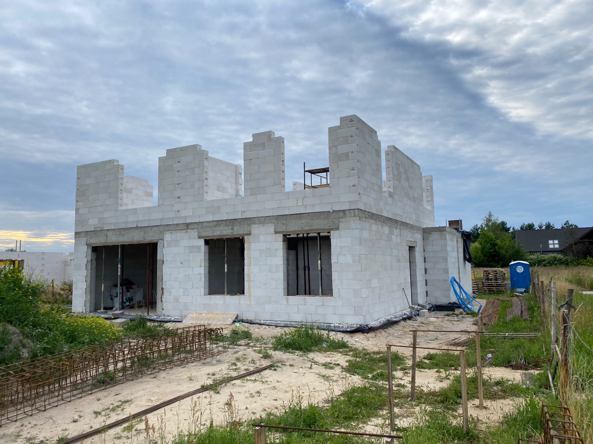 Wznoszenie ścian parterowego domu z białych bloczków, widoczna konstrukcja zbrojeniowa na gruncie i przenośna toaleta w tle.