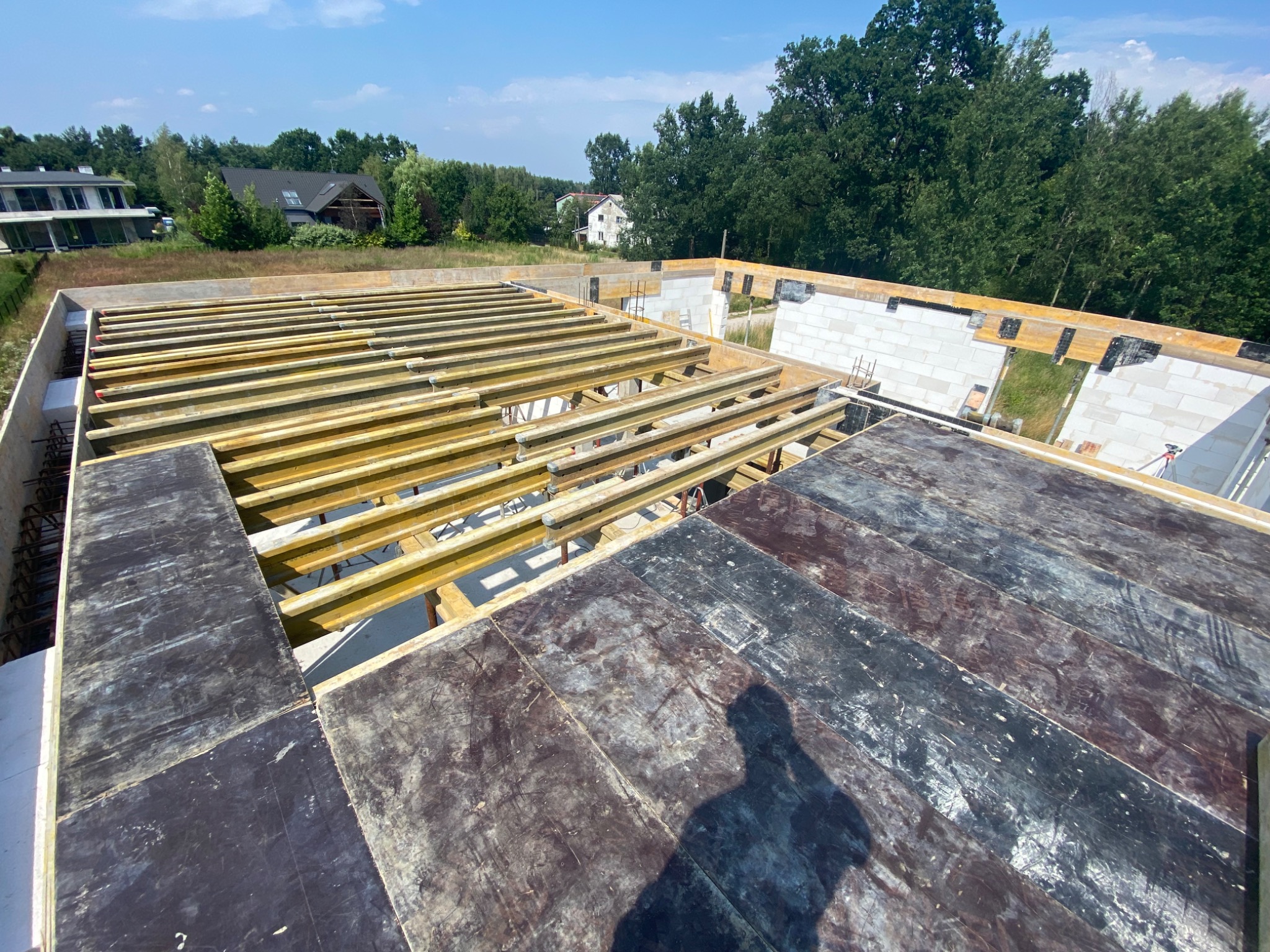 Wysoki kąt widzenia na fundamenty domu w trakcie budowy, widoczne szalunki i zbrojenie, tło z zielenią i domami.