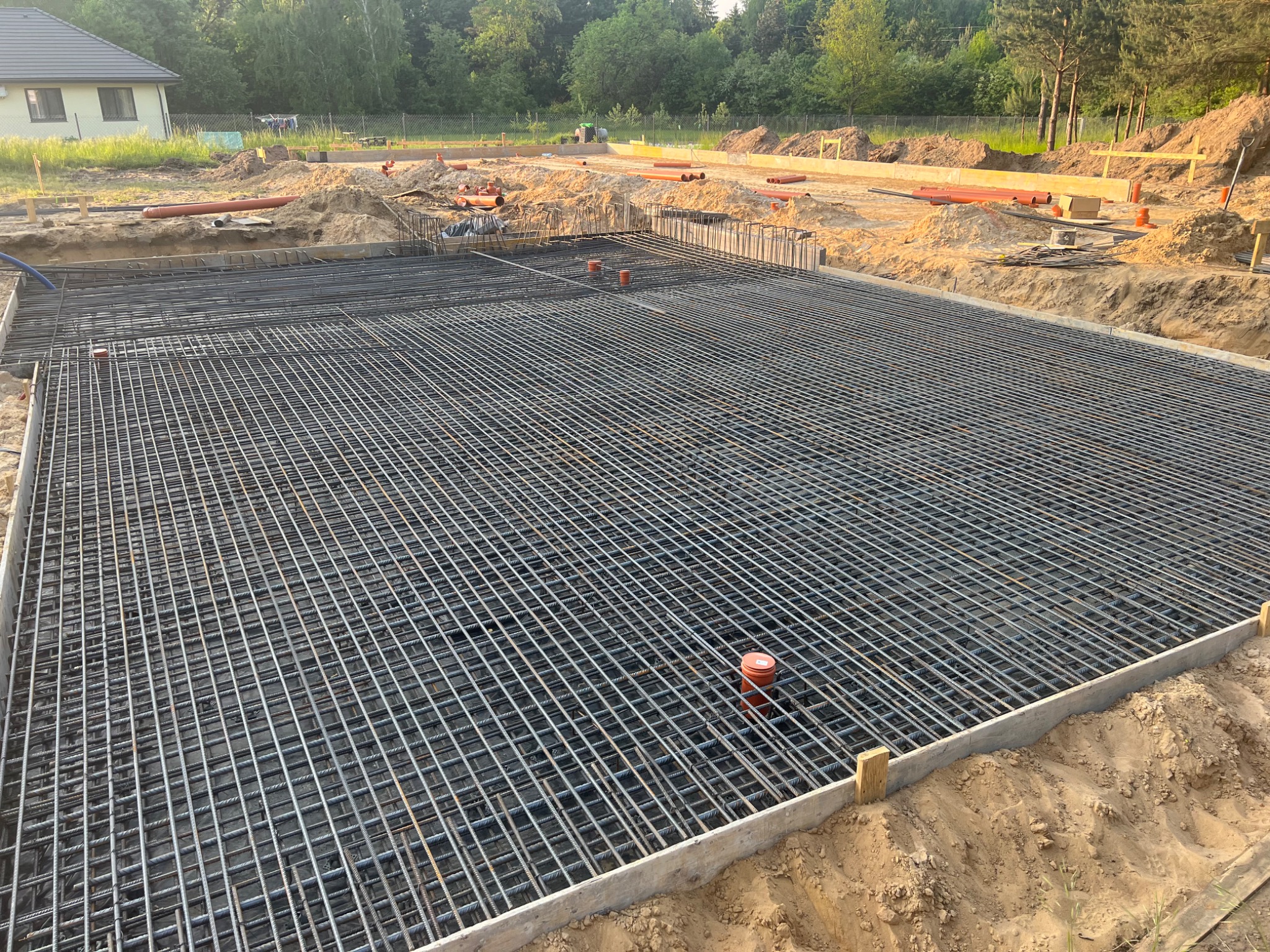 Siatka zbrojeniowa ułożona w szalunku fundamentowym, widoczne wystające rury kanalizacyjne, w tle dom w budowie i teren budowy z wykopami.