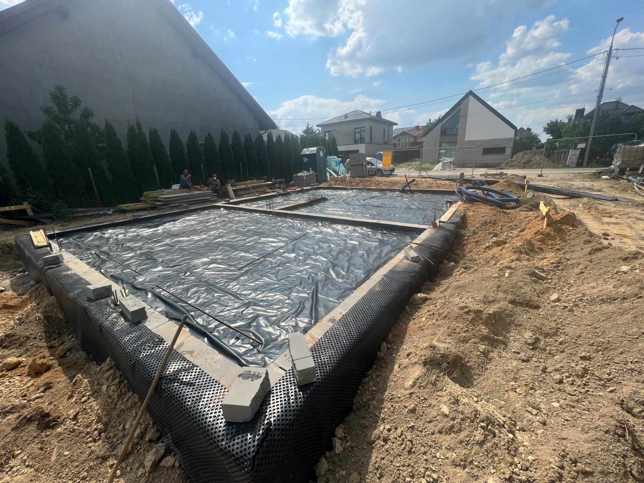 Przygotowany fundament pod budowę domu, z izolacją pionową i folią hydroizolacyjną, zbrojenie widoczne, teren budowy w słoneczny dzień.