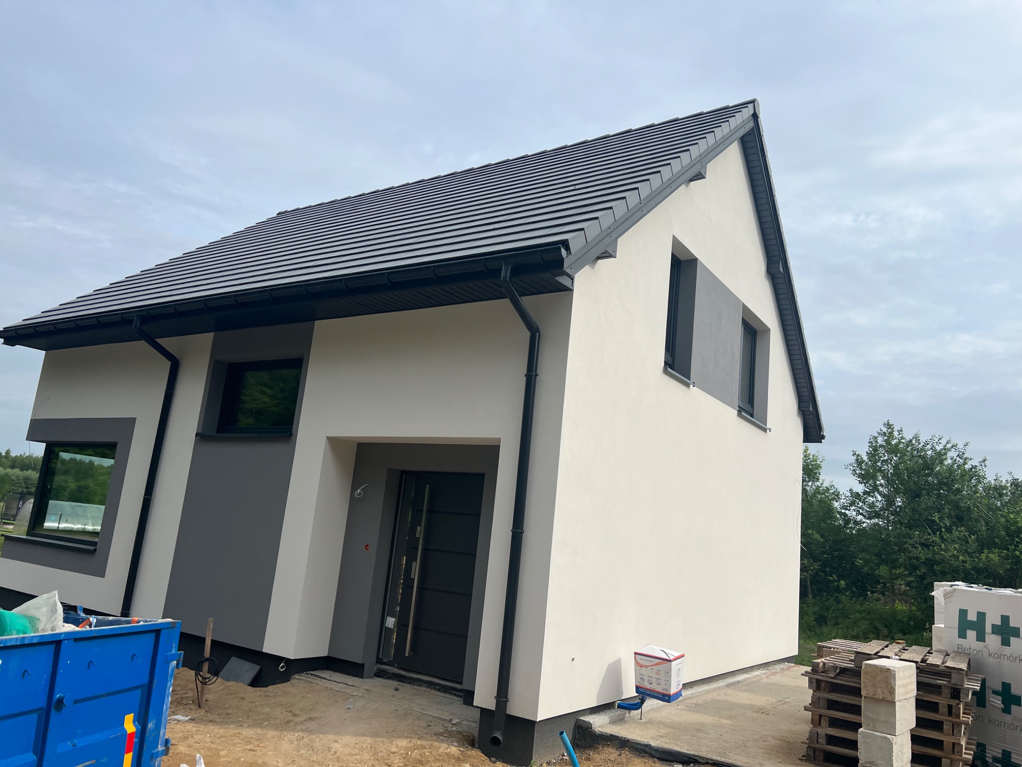 Nowoczesny dom jednorodzinny z szarą dachówką i jasną elewacją, widoczny kontener budowlany i palety z bloczkami, sugerujące trwające prace wykończeniowe.