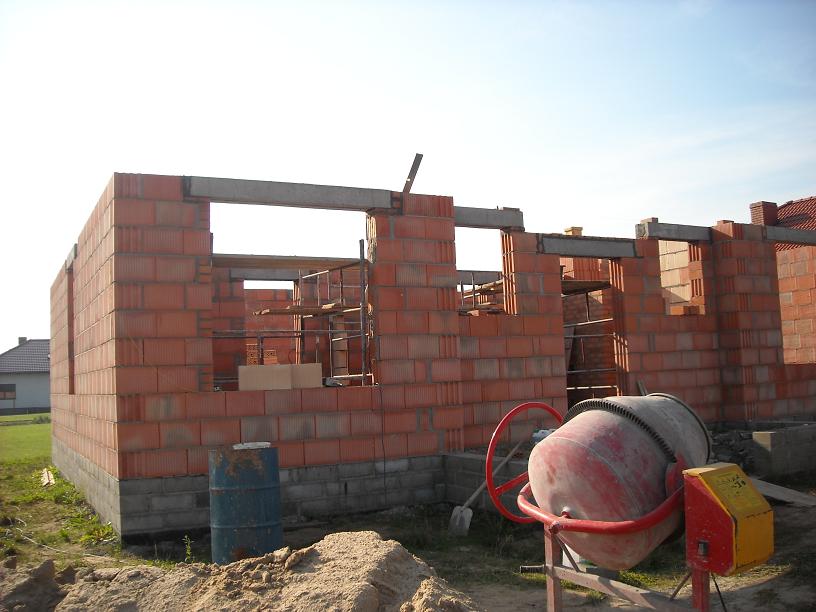 Wznoszenie ścian z czerwonej cegły na fundamencie z betonowych bloczków, widoczna betoniarka i rusztowanie wewnątrz budynku w stanie surowym otwartym.