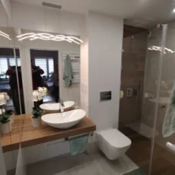 Nowoczesna łazienka z drewnianym blatem pod umywalką, białym sedesem i szklaną kabiną prysznicową z drewnianą ścianą w tle, oświetlona podwieszaną lampą w kształcie fal