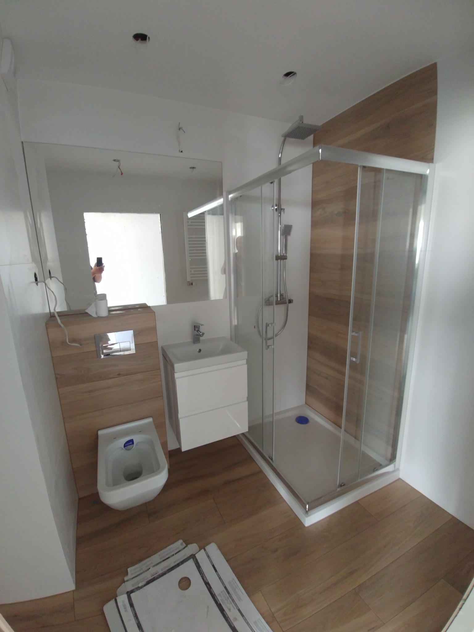 Nowoczesna łazienka z kabiną prysznicową, wiszącą miską WC zabudowaną w drewnopodobnej obudowie, umywalką na szafce i drewnopodobną okładziną ściany w kabinie prysznicowej. Na podłodze panele...