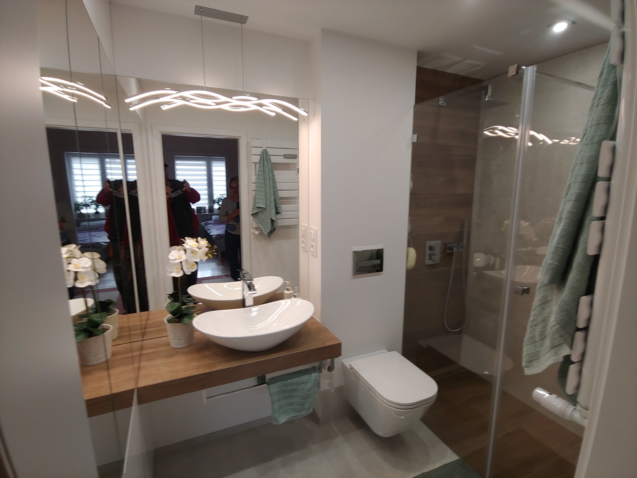 Nowoczesna łazienka z drewnianym blatem pod umywalką, białym sedesem i szklaną kabiną prysznicową z drewnianą ścianą w tle, oświetlona podwieszaną lampą w kształcie fal