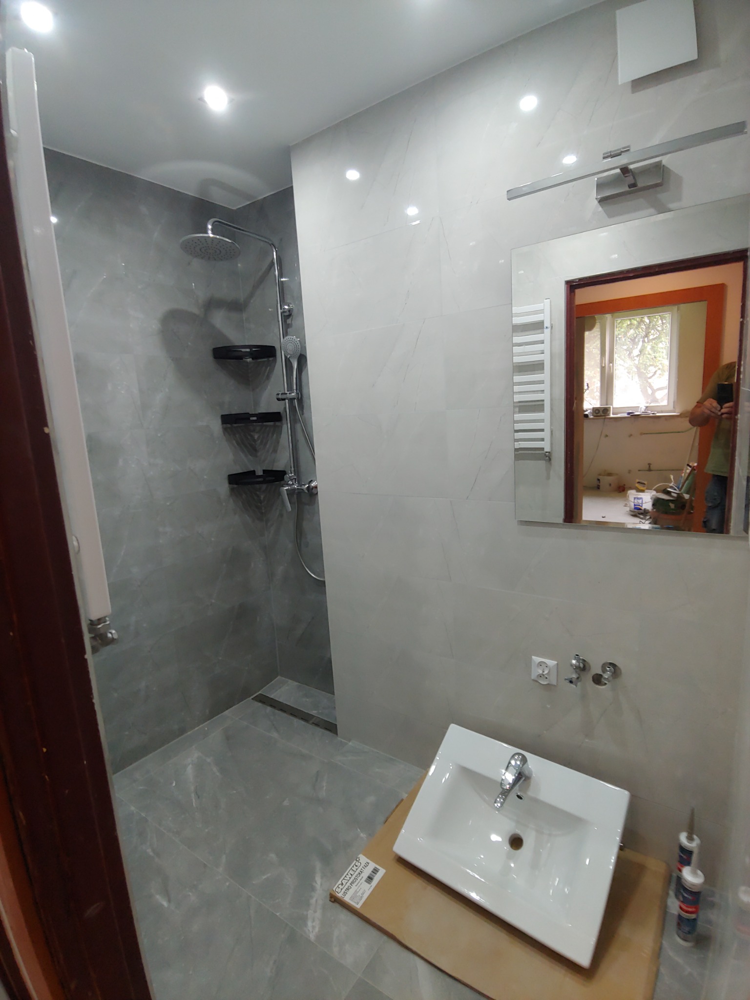 Szara łazienka z kabiną prysznicową typu walk-in, umywalką na kartonie, i lustrem odbijającym remontowane pomieszczenie.