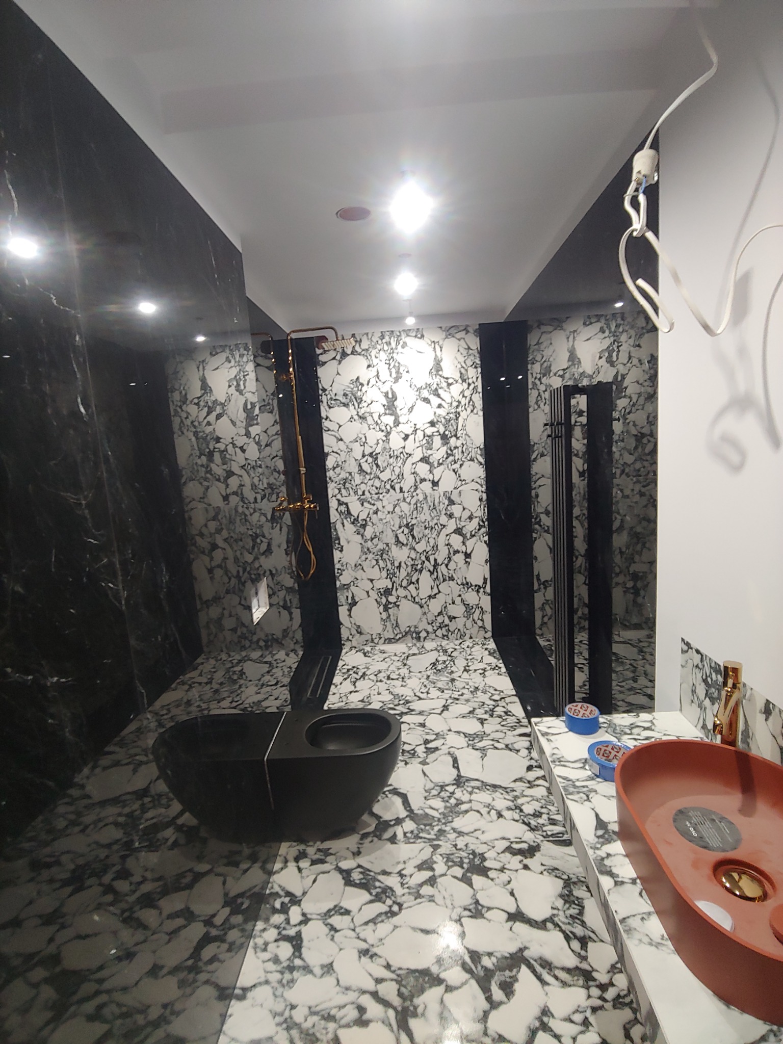 Nowoczesna łazienka z czarno-białymi marmurowymi płytkami, czarną umywalką nablatową i miedzianą armaturą, widoczne puszki z silikonem na blacie.