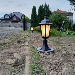 Posmar - Dwa czarne, oświetlone lampiony ogrodowe stojące w rzędzie na ziemi, z widocznymi domami w tle.