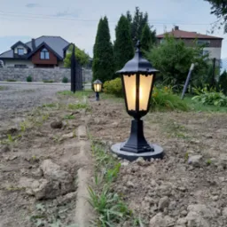 Dwa czarne, oświetlone lampiony ogrodowe stojące w rzędzie na ziemi, z widocznymi domami w tle.