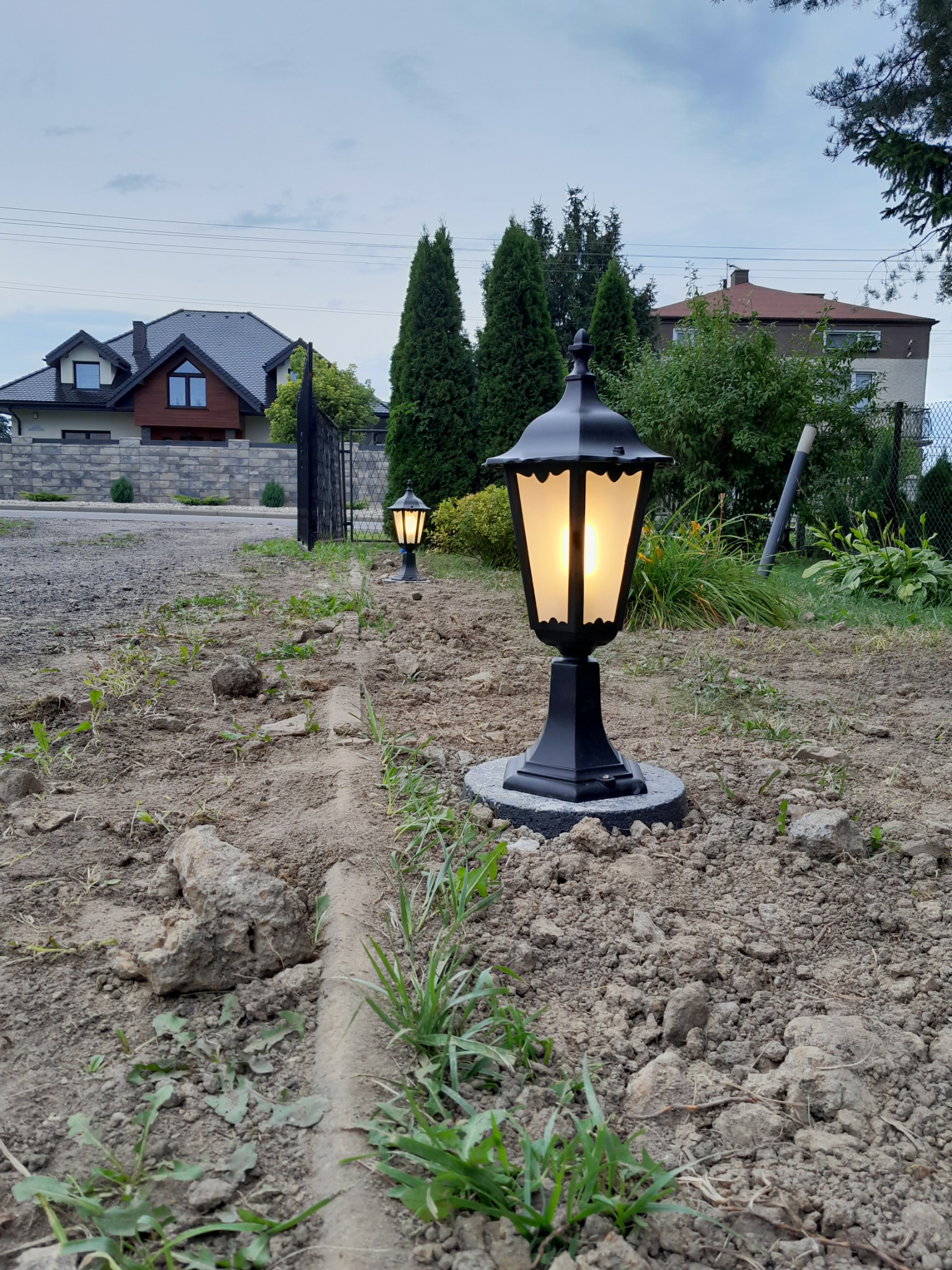 Dwa czarne, oświetlone lampiony ogrodowe stojące w rzędzie na ziemi, z widocznymi domami w tle.