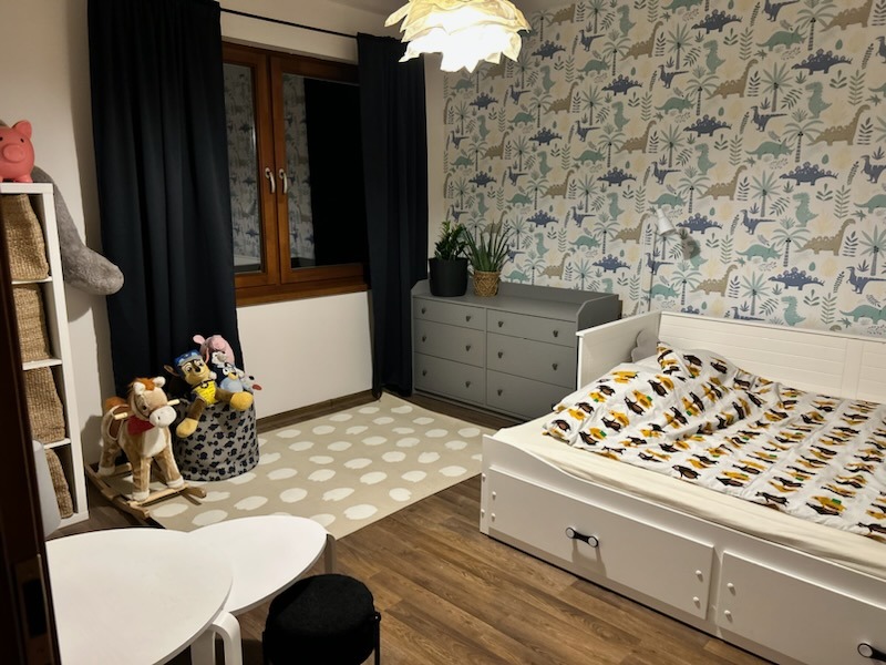 Pokój dziecięcy z dinozaurową tapetą, białym łóżkiem z szufladami, szarą komodą i ciemnymi zasłonami przy oknie.