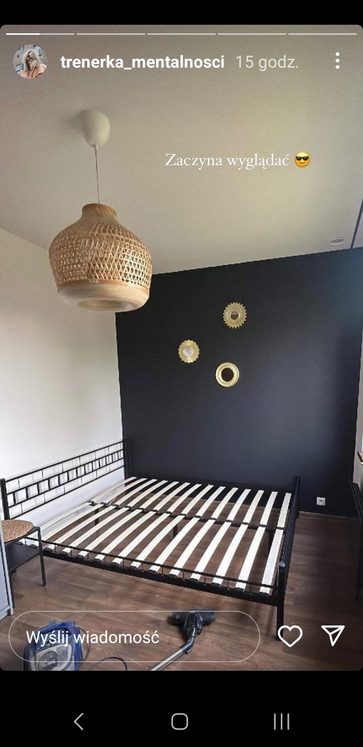 Pokój z czarną ścianą, metalową ramą łóżka z drewnianymi listwami, plecioną lampą i złotymi dekoracjami ściennymi, odkurzacz na podłodze.