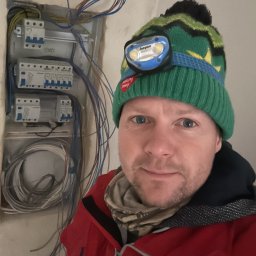 REAL-ELECTRIK ŁUKASZ BEDNAREK - Elektryk w czapce z latarką czołową, pozuje na tle otwartej skrzynki z bezpiecznikami i przewodami w trakcie prac instalacyjnych w surowym wnętrzu.