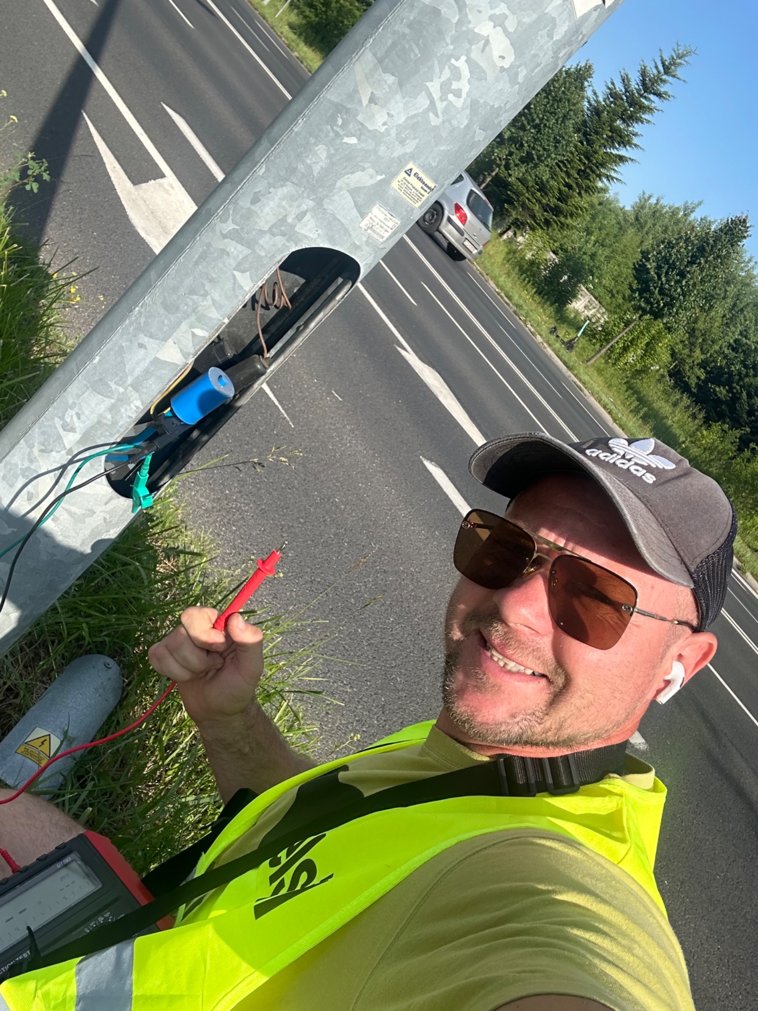Elektryk w kamizelce odblaskowej, z miernikiem w ręku, uśmiechnięty, podczas pomiarów w otwartej skrzynce słupa oświetleniowego przy drodze.