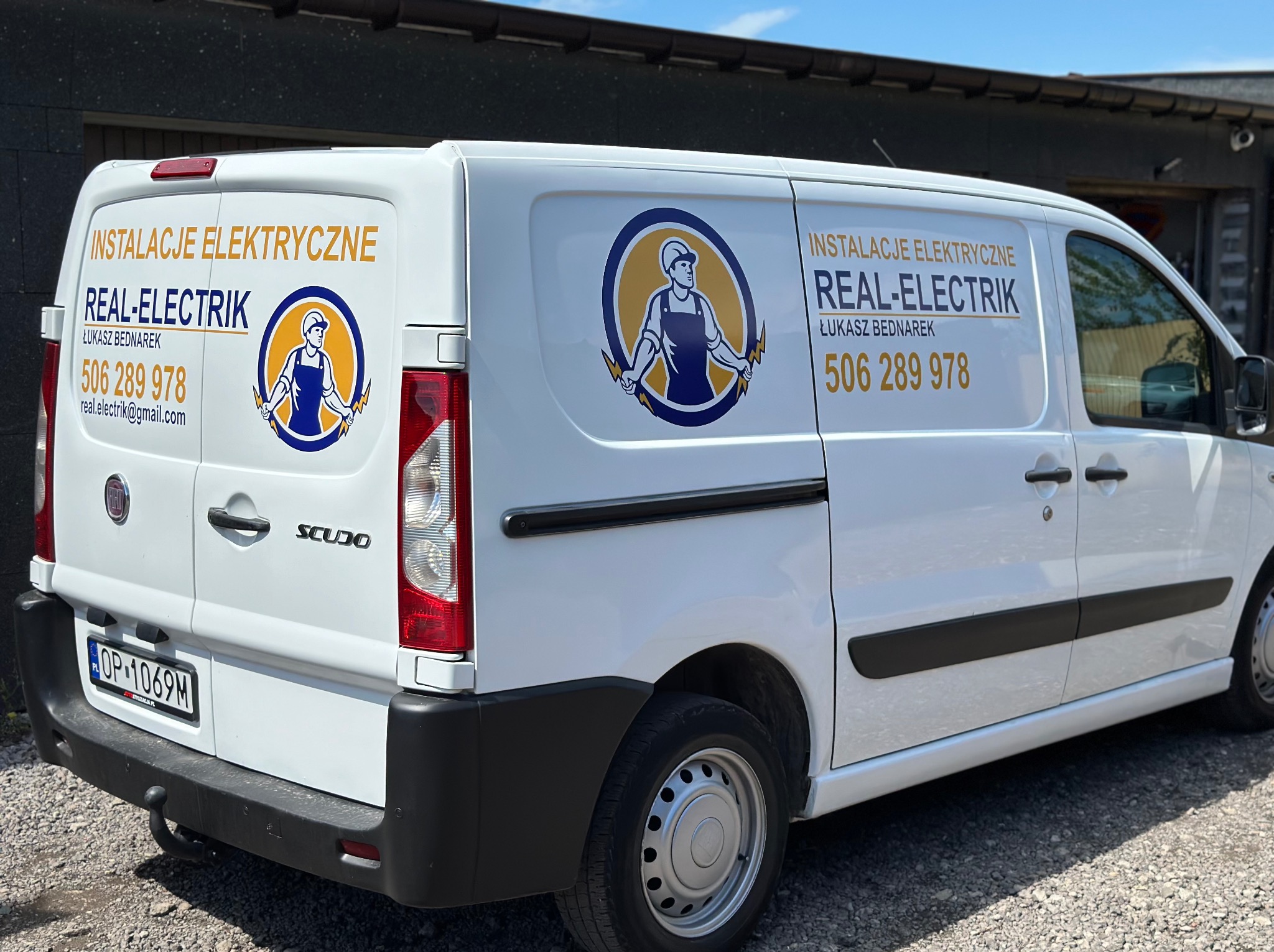 Biały samochód dostawczy firmy Real-Elektrik z numerem telefonu i logo przedstawiającym elektryka trzymającego błyskawice, zaparkowany na żwirowym podłożu.