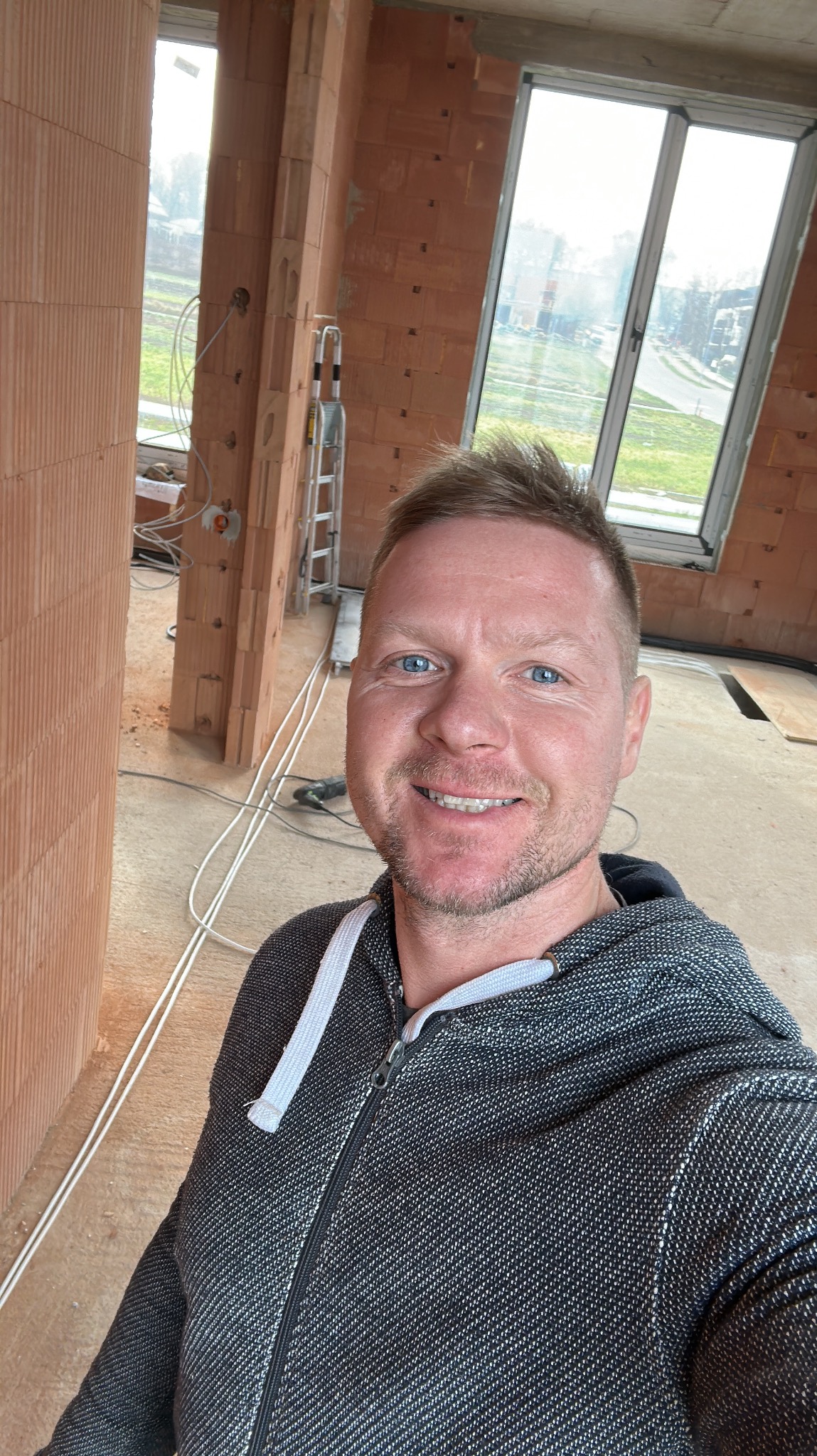 Selfie elektryka na tle surowych ścian z cegły ceramicznej w trakcie instalacji elektrycznej, widoczne kable i puszki instalacyjne, drabina aluminiowa w tle.