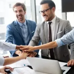 Radosny zespół biznesowy z rękami złożonymi w piramidę nad biurkiem w nowoczesnym biurze, wyrażający jedność i sukces.