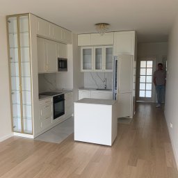 M.A.K.S. Alona Tkachuk - Jasna kuchnia w mieszkaniu z białymi meblami, złotymi akcentami i marmurowym blatem, widok na korytarz z osobą w tle.