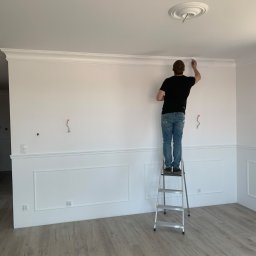 M.A.K.S. Alona Tkachuk - Mężczyzna maluje białą listwę dekoracyjną pod sufitem, stojąc na aluminiowej drabinie w jasnym pomieszczeniu z panelami podłogowymi, widoczne wystające kable ze ściany.