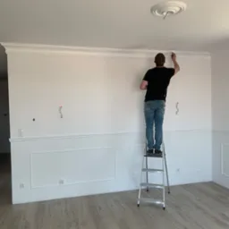 Mężczyzna maluje białą listwę dekoracyjną pod sufitem, stojąc na aluminiowej drabinie w jasnym pomieszczeniu z panelami podłogowymi, widoczne wystające kable ze ściany.