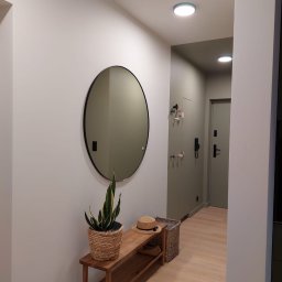 M.A.K.S. Alona Tkachuk - Nowoczesny przedpokój w odcieniach szarości i beżu z okrągłym lustrem, drewnianą ławką z plecionym koszem i rośliną w wiklinowej doniczce.