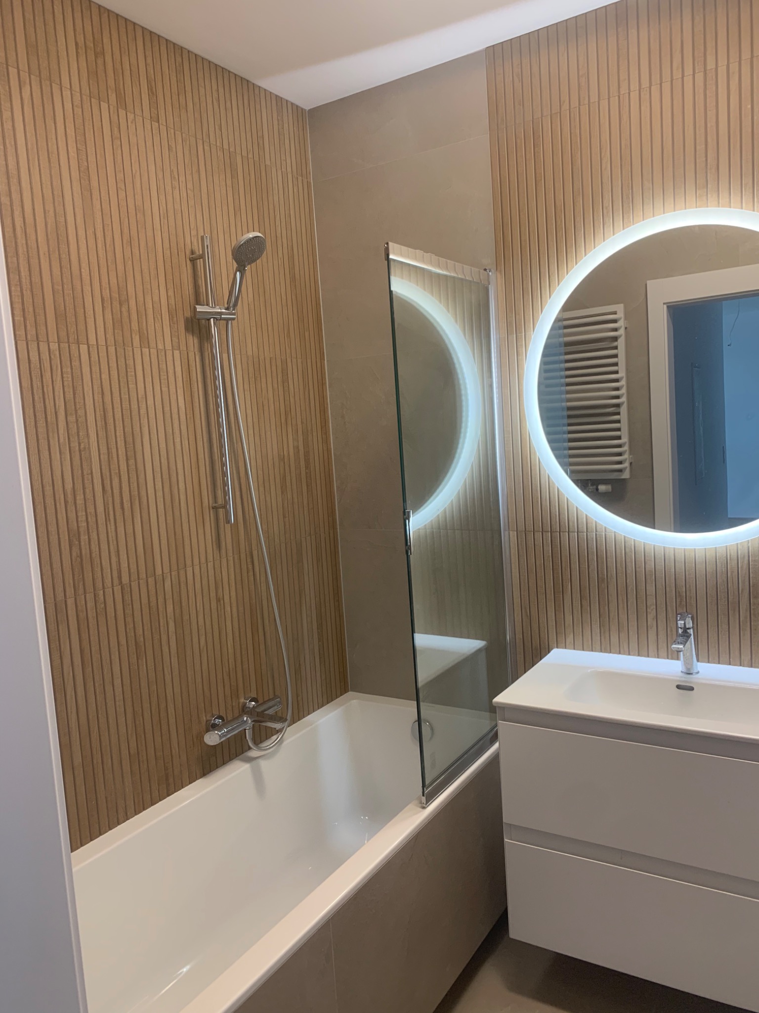 Nowoczesna łazienka z wanną, szklanym parawanem, umywalką z szafką i okrągłym lustrem z podświetleniem LED, ściany wyłożone płytkami imitującymi drewno.