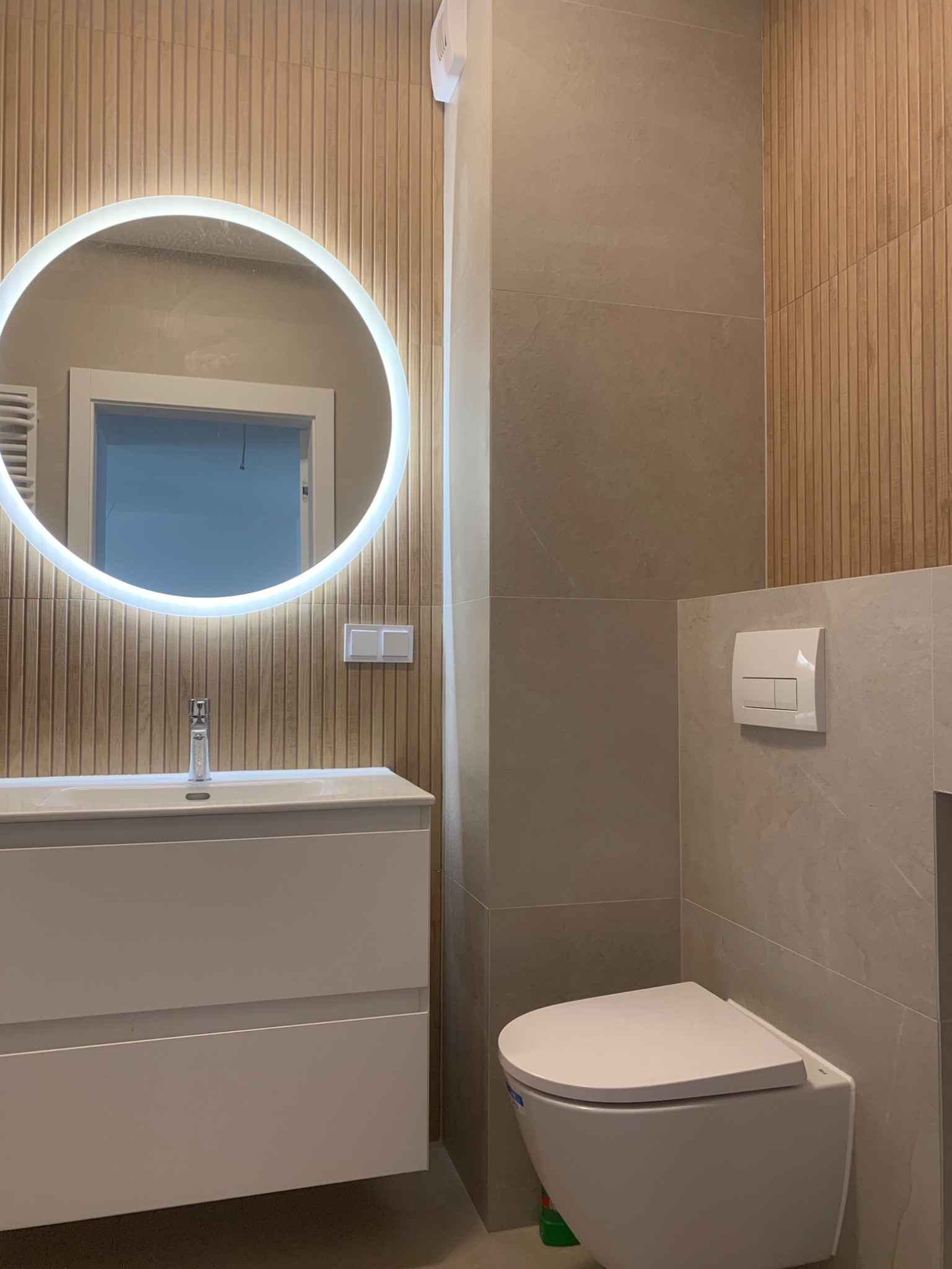 Nowoczesna łazienka z podświetlanym okrągłym lustrem, białą szafką pod umywalką, wiszącą toaletą i płytkami imitującymi drewno na ścianie.