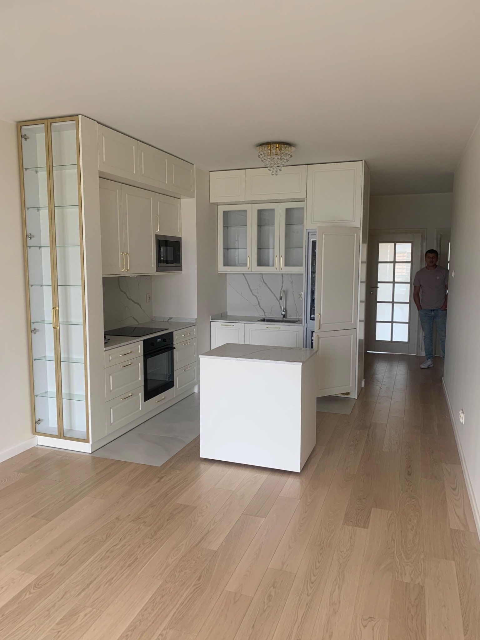 Jasna kuchnia w mieszkaniu z białymi meblami, złotymi akcentami i marmurowym blatem, widok na korytarz z osobą w tle.