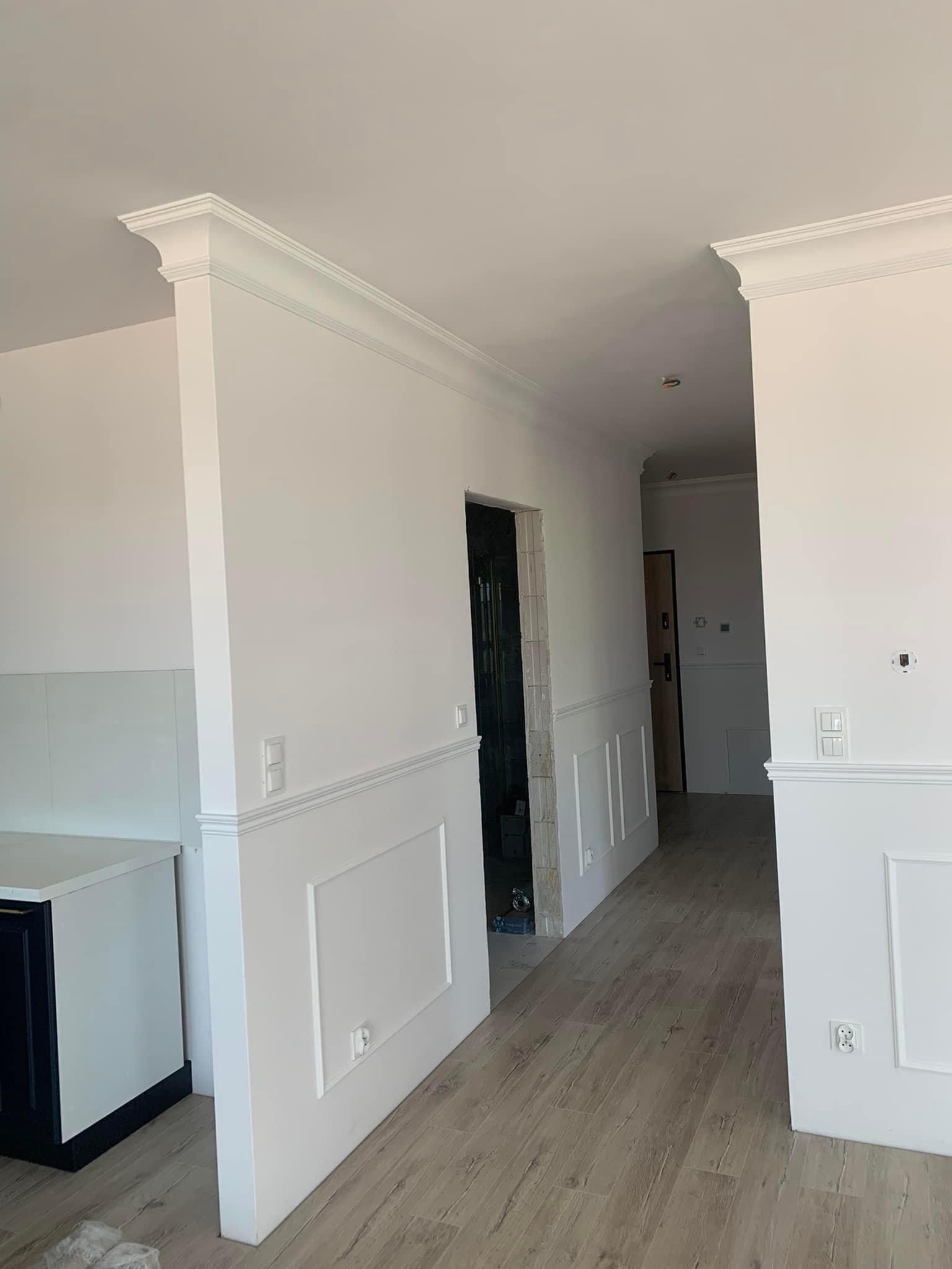 Białe ściany z dekoracyjnymi panelami i listwami przypodłogowymi w jasnym wnętrzu, widok na korytarz z drewnianą podłogą, gniazdka elektryczne zamontowane na ścianach.