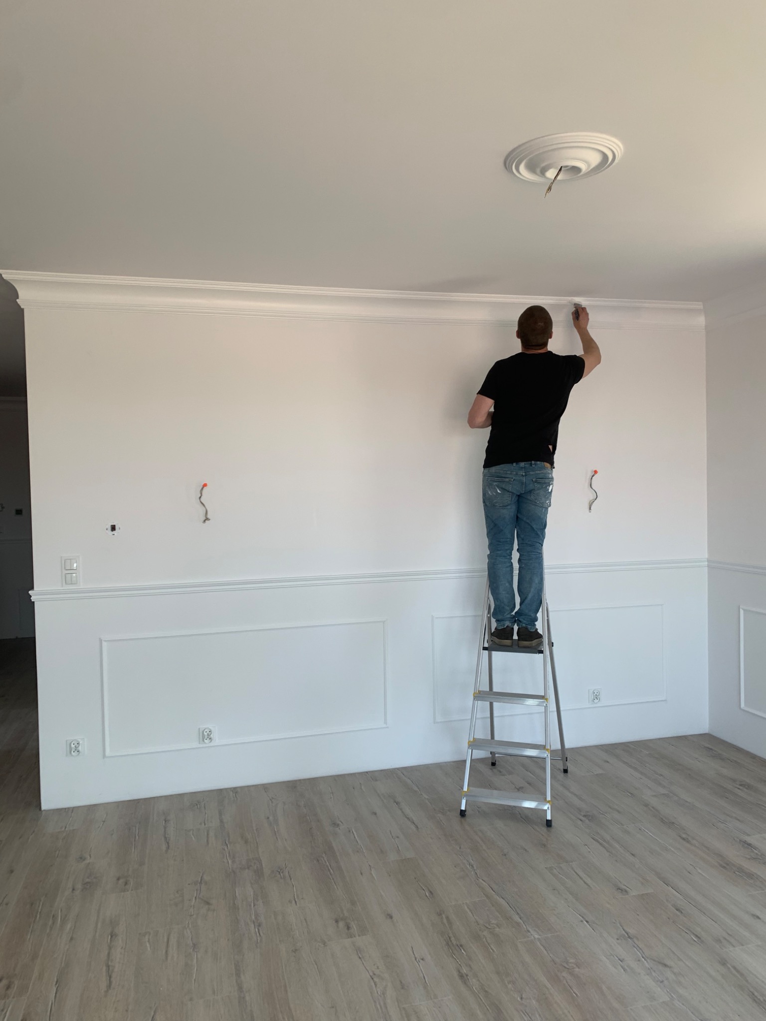 Mężczyzna maluje białą listwę dekoracyjną pod sufitem, stojąc na aluminiowej drabinie w jasnym pomieszczeniu z panelami podłogowymi, widoczne wystające kable ze ściany.