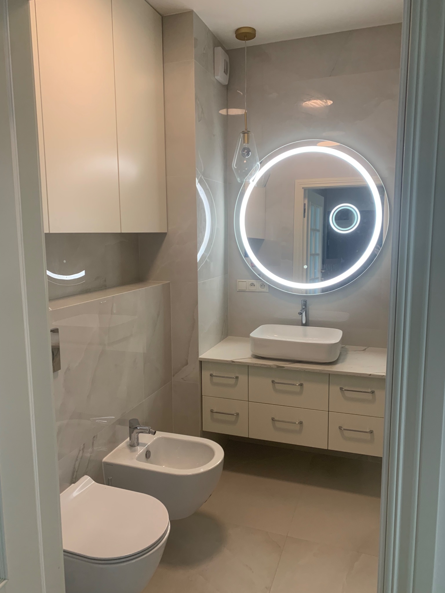 Nowoczesna łazienka w Gdańsku z podświetlanym okrągłym lustrem, białą umywalką nablatową, szafką z szufladami i wiszącą toaletą z bidetem.
