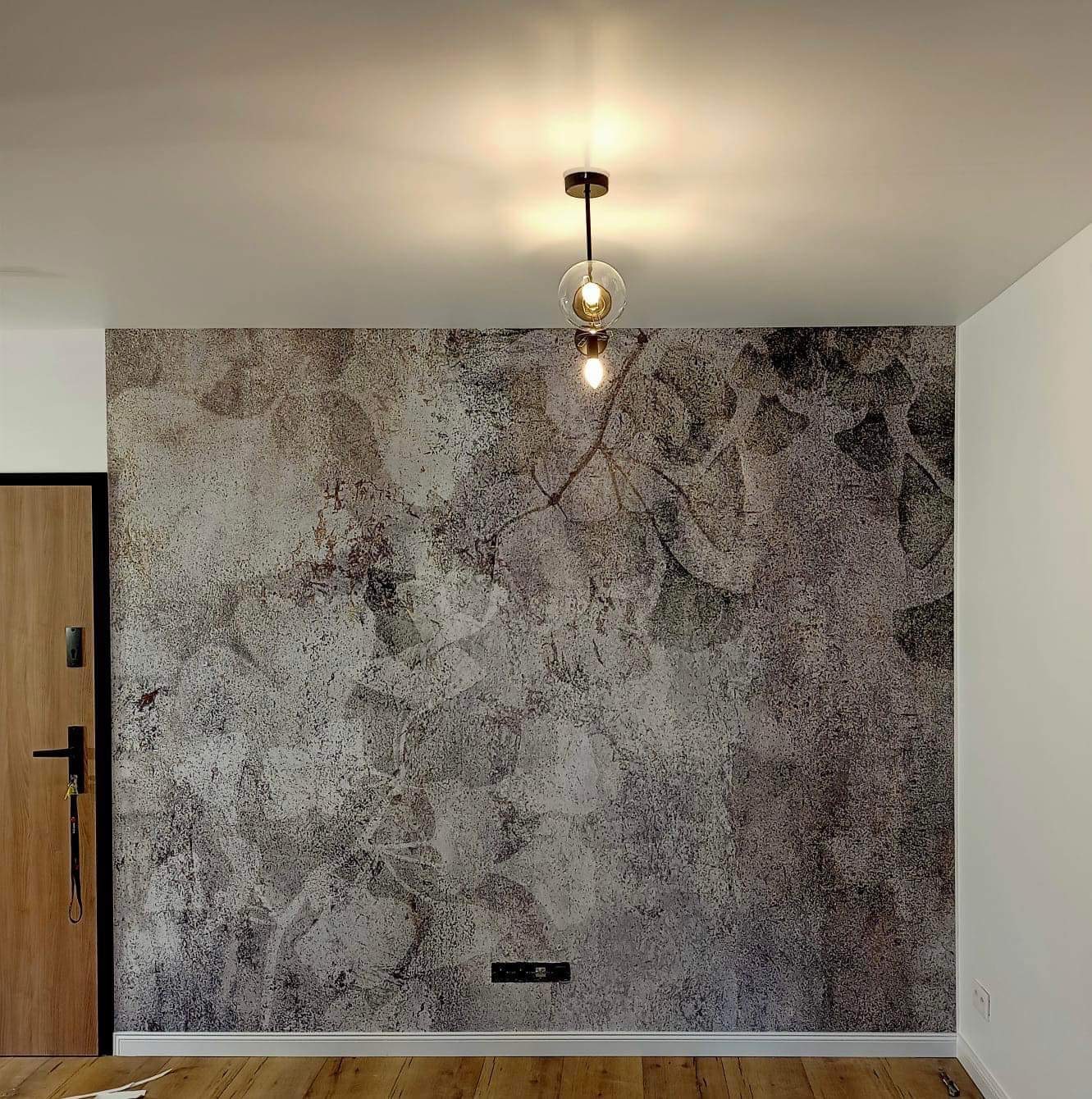 Ściana pokryta dekoracyjną tapetą imitującą strukturę kamienia z motywem roślinnym, oświetlona przez nowoczesną lampę wiszącą z dwoma szklanymi kloszami, drewniana podłoga i drzwi.