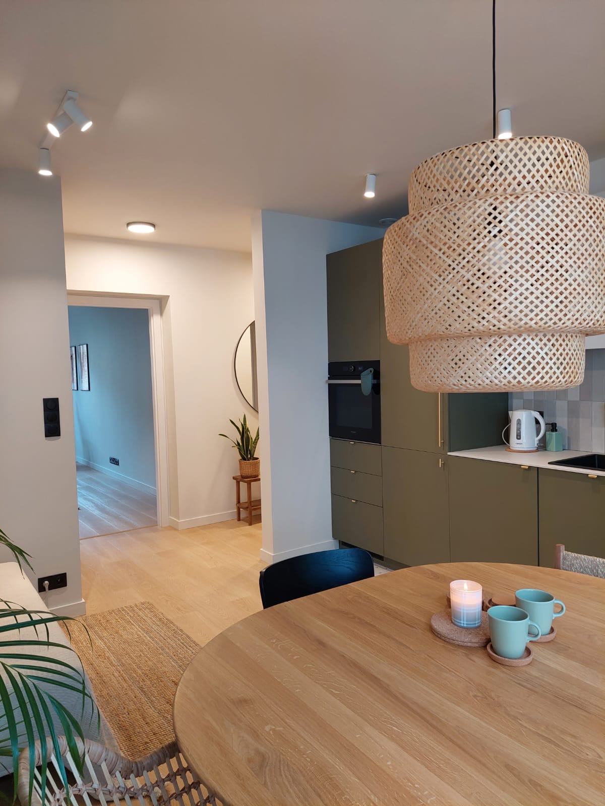 Jasne wnętrze mieszkania z drewnianym stołem, plecioną lampą wiszącą i zieloną zabudową kuchenną z piekarnikiem