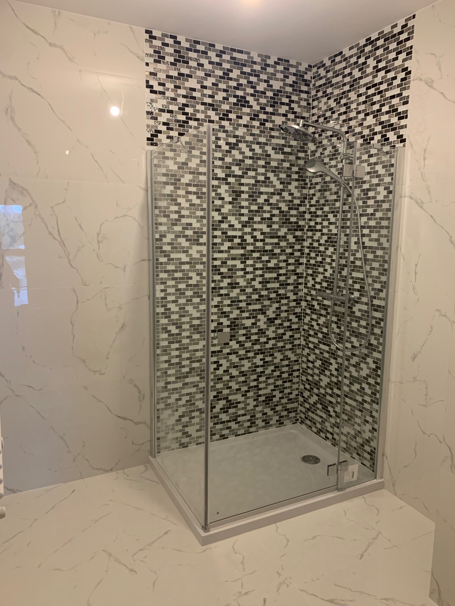 Kabina prysznicowa narożna z przezroczystego szkła, wykończona mozaiką w odcieniach szarości i bieli, na ścianach i podłodze białe płytki imitujące marmur.