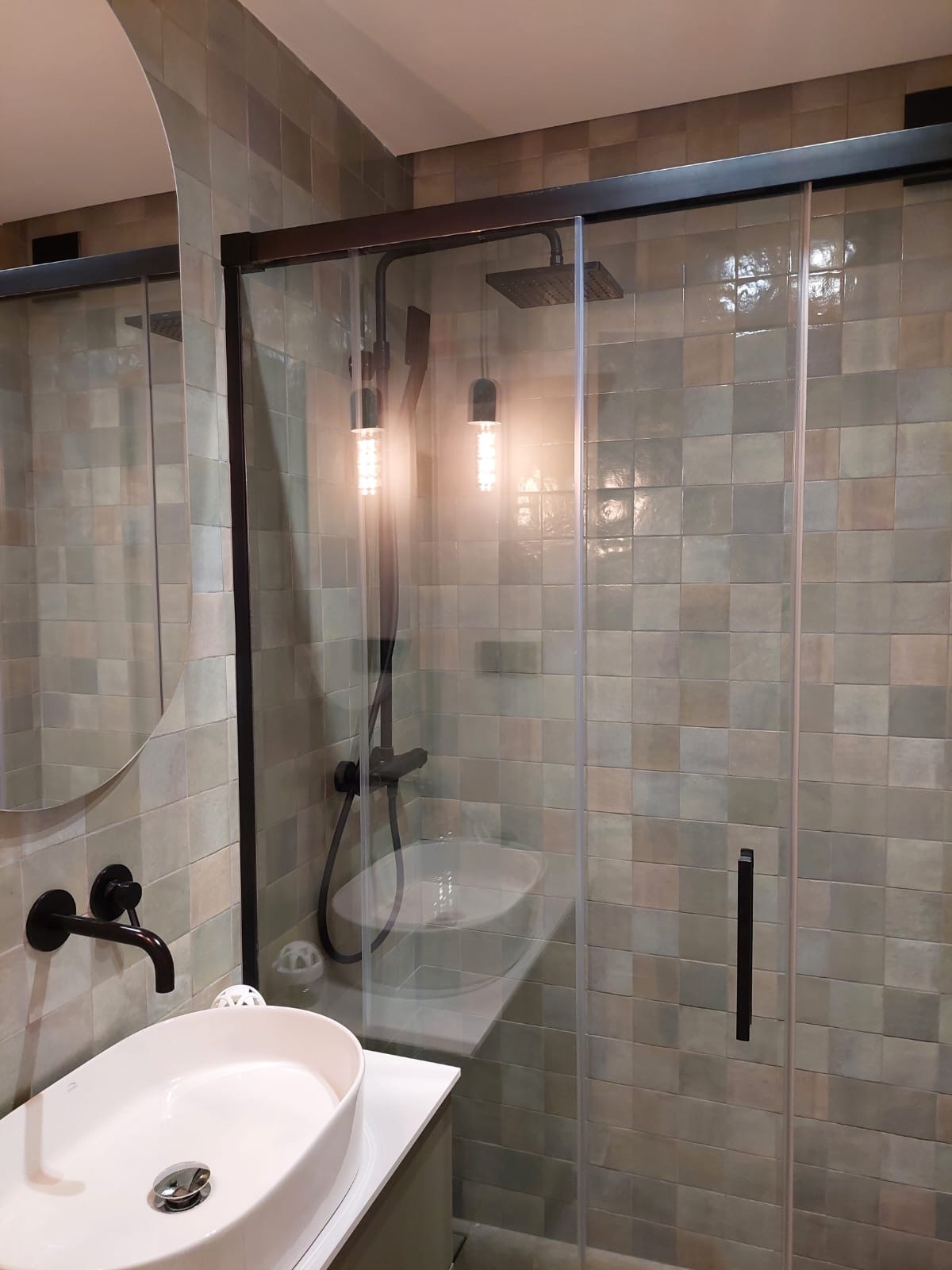 Nowoczesna łazienka z kabiną prysznicową typu walk-in, wykończona kwadratowymi płytkami w odcieniach beżu i zieleni, z czarną armaturą i okrągłym lustrem.