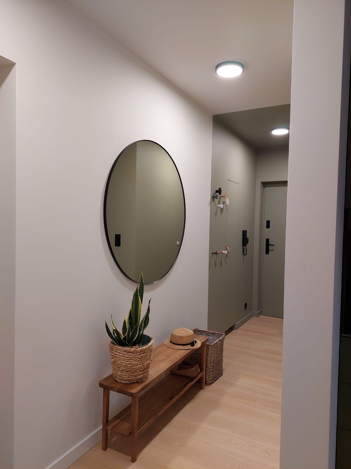 Nowoczesny przedpokój w odcieniach szarości i beżu z okrągłym lustrem, drewnianą ławką z plecionym koszem i rośliną w wiklinowej doniczce.