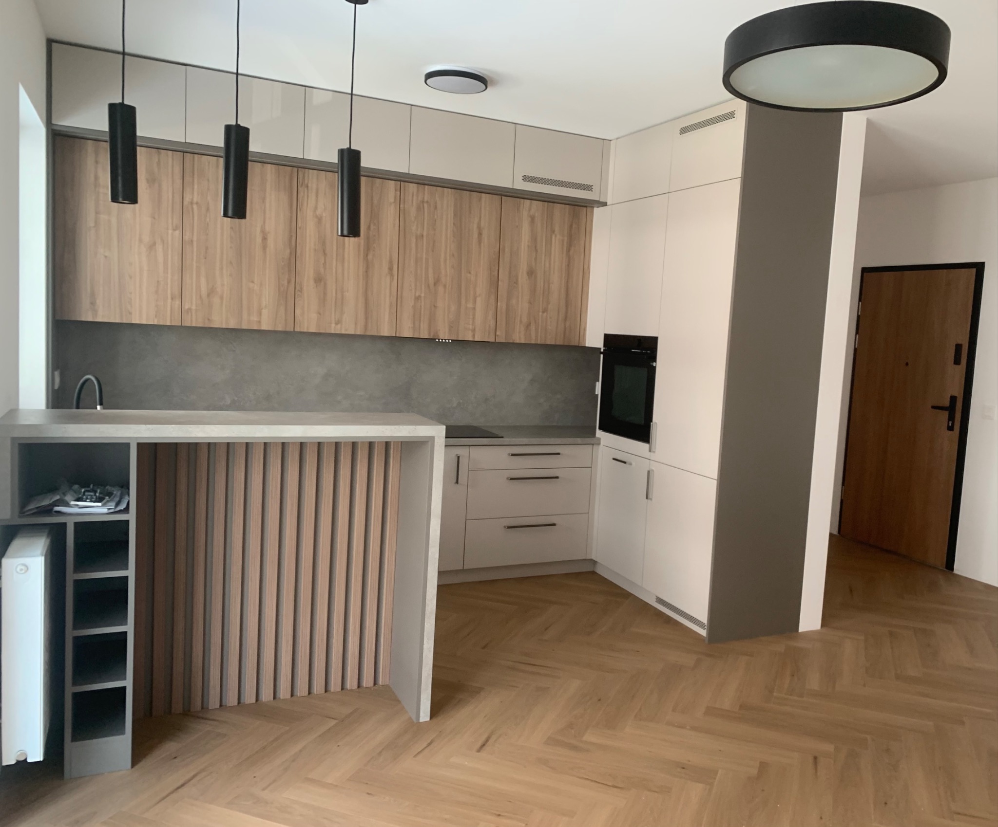 Nowoczesna kuchnia z drewnianymi i białymi szafkami, szarym blatem, wyspą z pionowymi listwami i parkietem ułożonym w jodełkę.