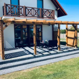 Drewniana konstrukcja zadaszenia tarasu przy domu z balkonem, meble ogrodowe na kostce brukowej i trawnik.