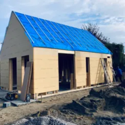 Budowa domu szkieletowego z niebieską folią na dachu, widoczne płyty konstrukcyjne, drabina oparta o ścianę, oraz kopiec żwiru na pierwszym planie.